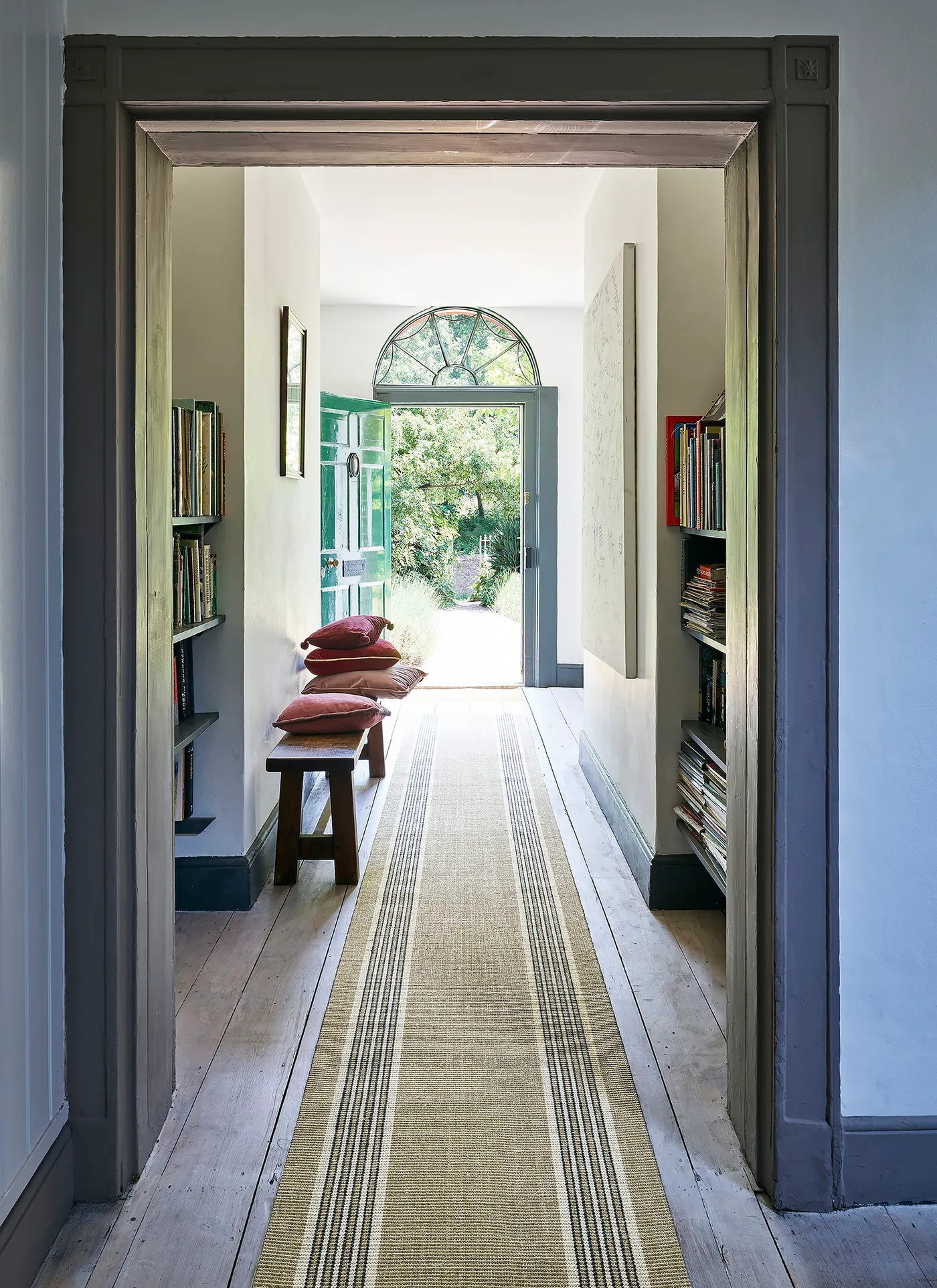 Long pale runner with dark striped borders between wood bench and bookshelves, leading to an open glazed arched door