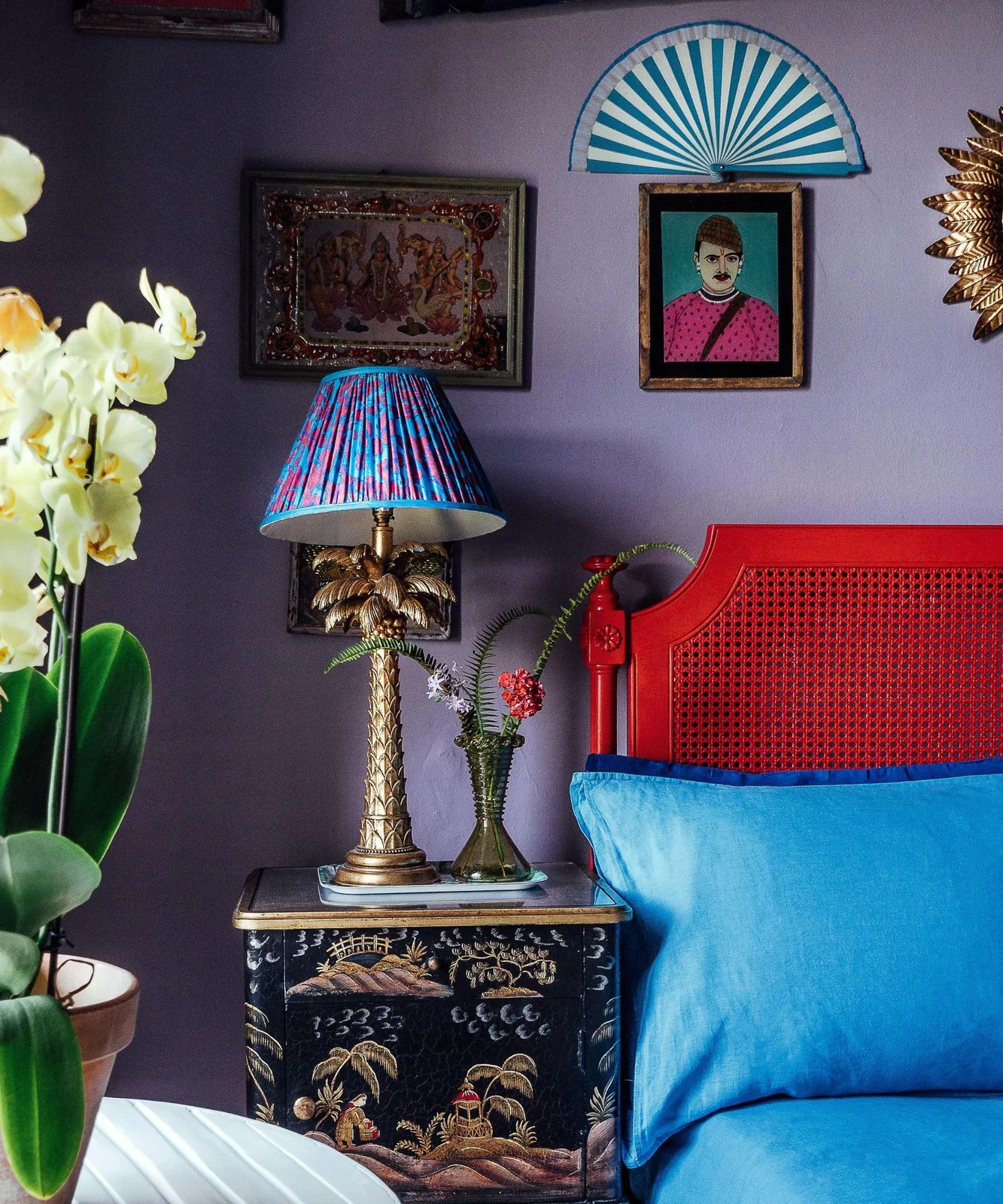 A red woven headboard sits against a purple wall beside framed portraits, a decorative fan and a palm-shaped lamp with a striped shade