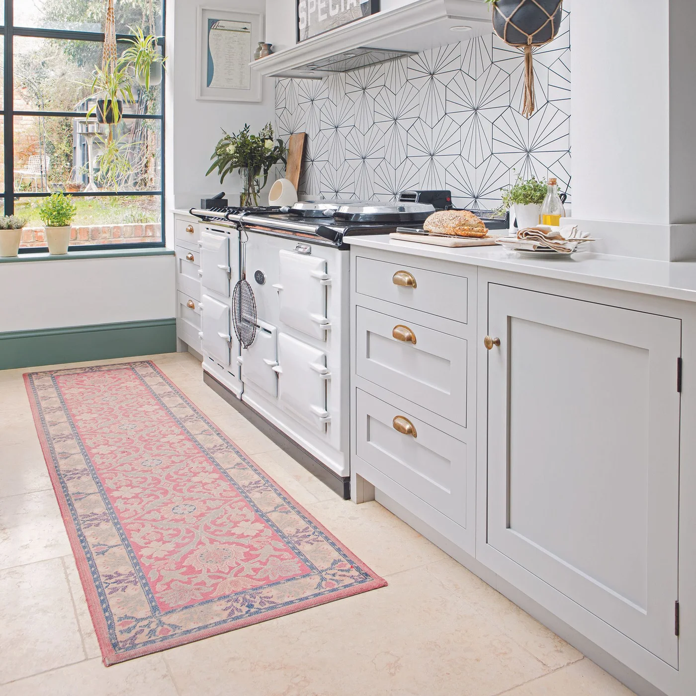 Pale gray cabinets with gold-toned cup handles sit beside a white range cooker beneath a geometric backsplash and above a pink patterned runner