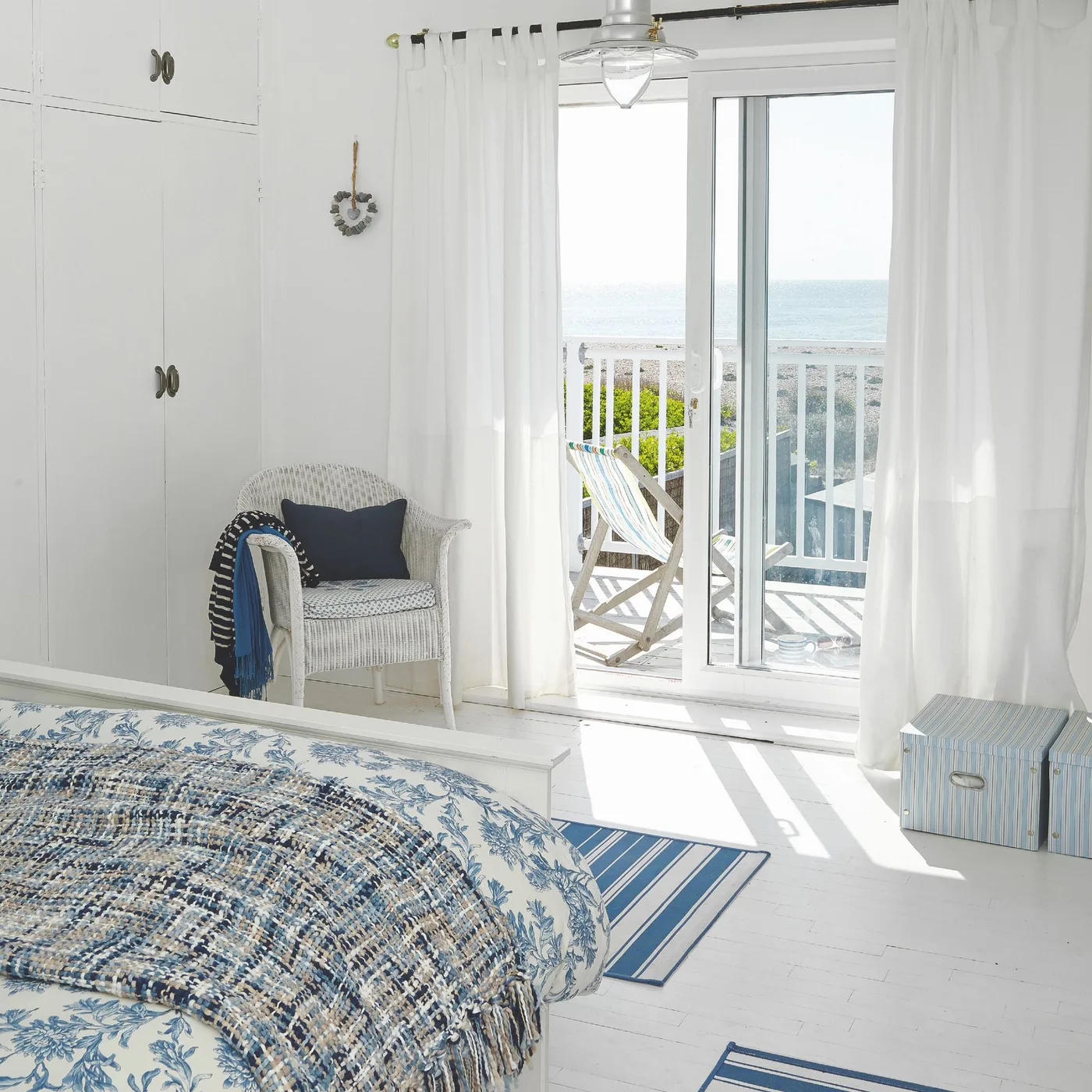 White coastal bedroom with blue patterned bedding, a woven chair, striped floor rugs and glazed balcony doors.