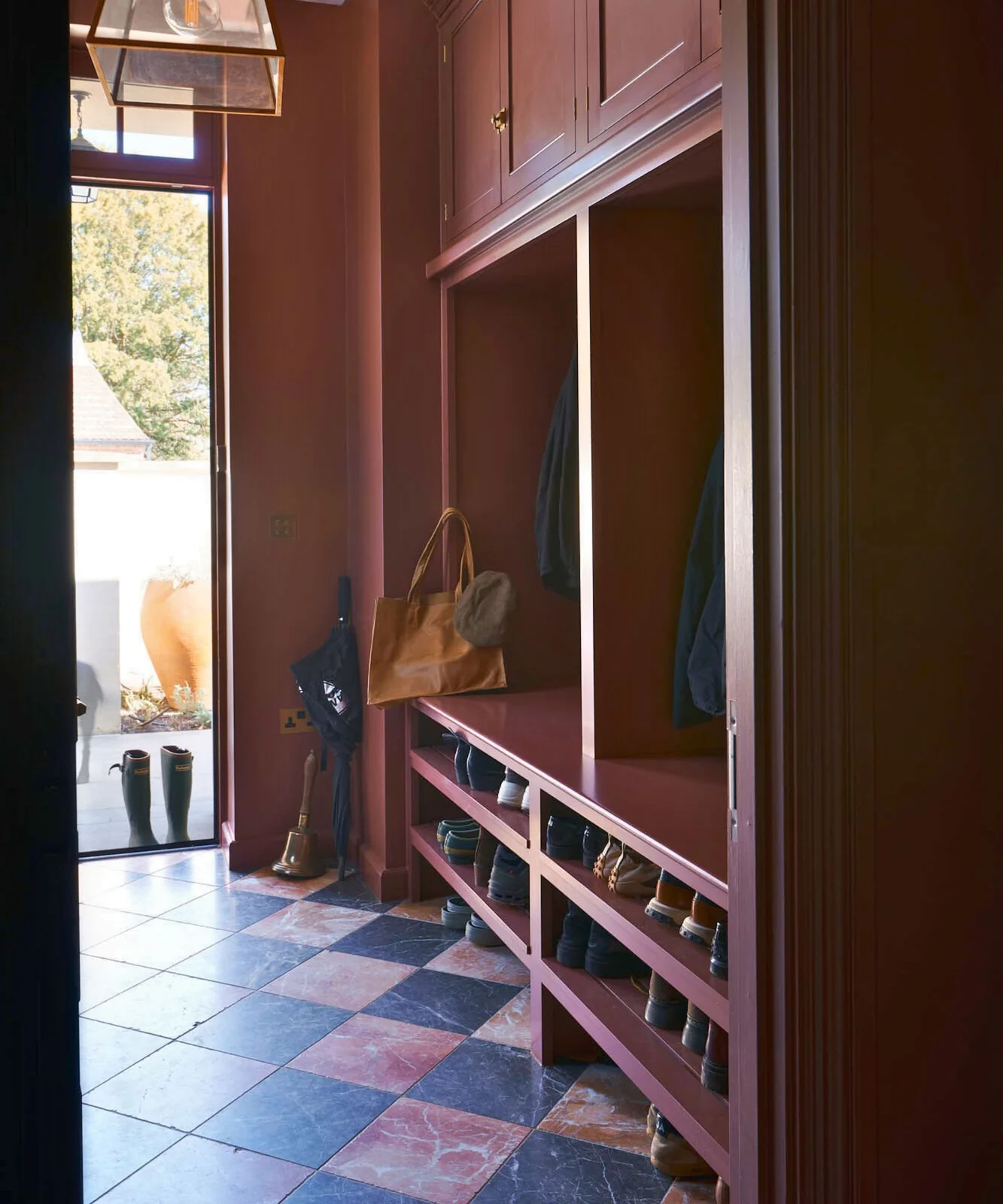 Burgundy framed cupboards and open coat bays stand above two rows of shoe shelves beside a dark glazed doorway and a checked tile floor