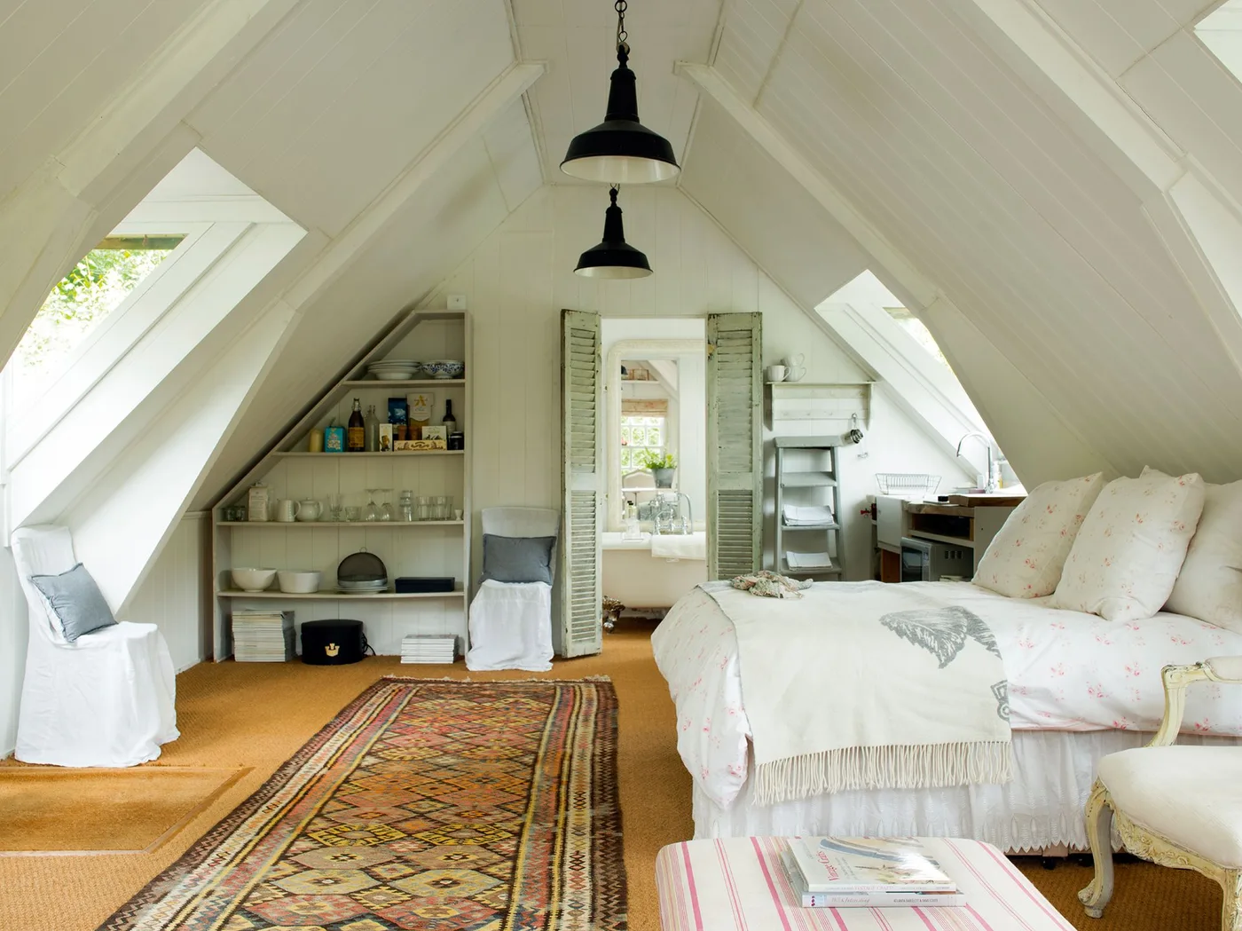 Pale attic bedroom with a bed beneath a sloping roof, open shelving, covered chairs, wooden floor and a long patterned rug.