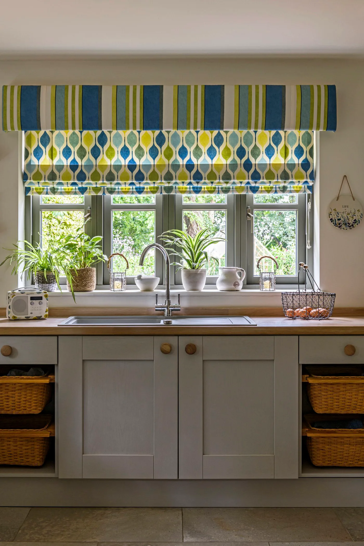 Cottage kitchen sink area with gray framed cabinets, woven storage baskets, wooden countertop and blue-and-yellow patterned blind beneath a striped valance.