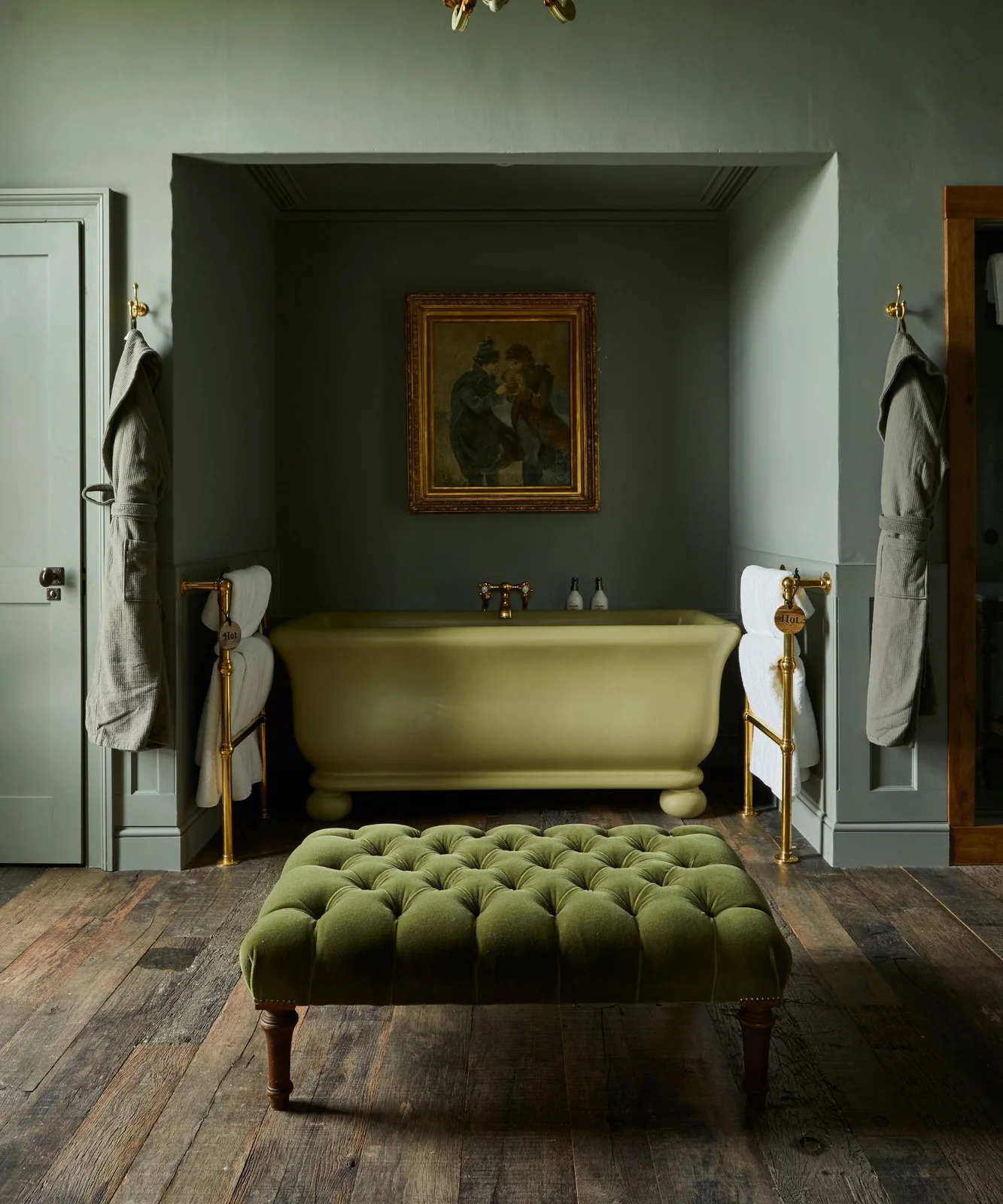 A green bath sits in a darker green alcove below framed artwork behind a tufted green ottoman and warm metal towel rails