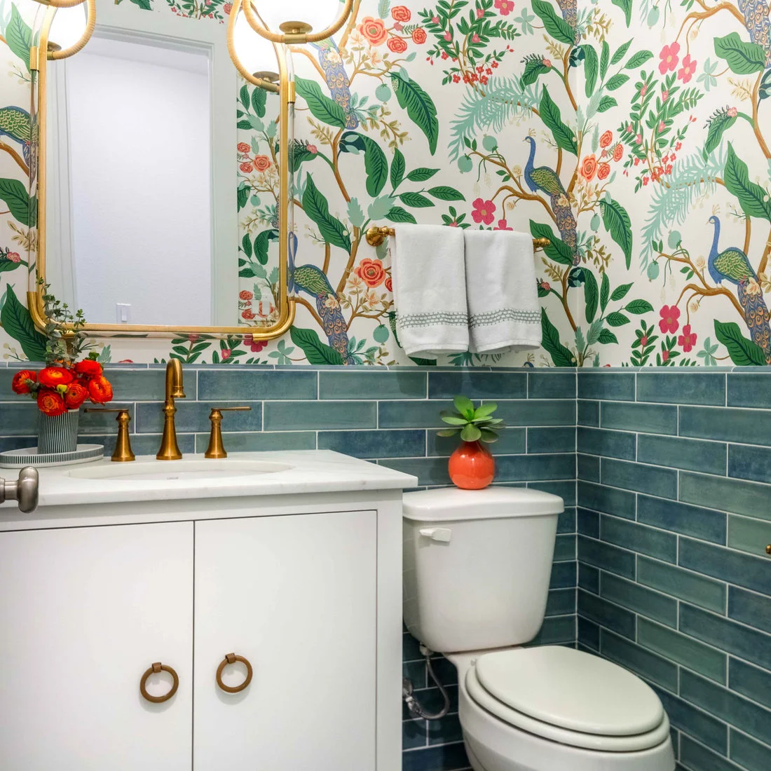 Peacock and floral wallpaper sits above blue-green rectangular tiles beside a white powder-room vanity, gold-colored mirror and fittings