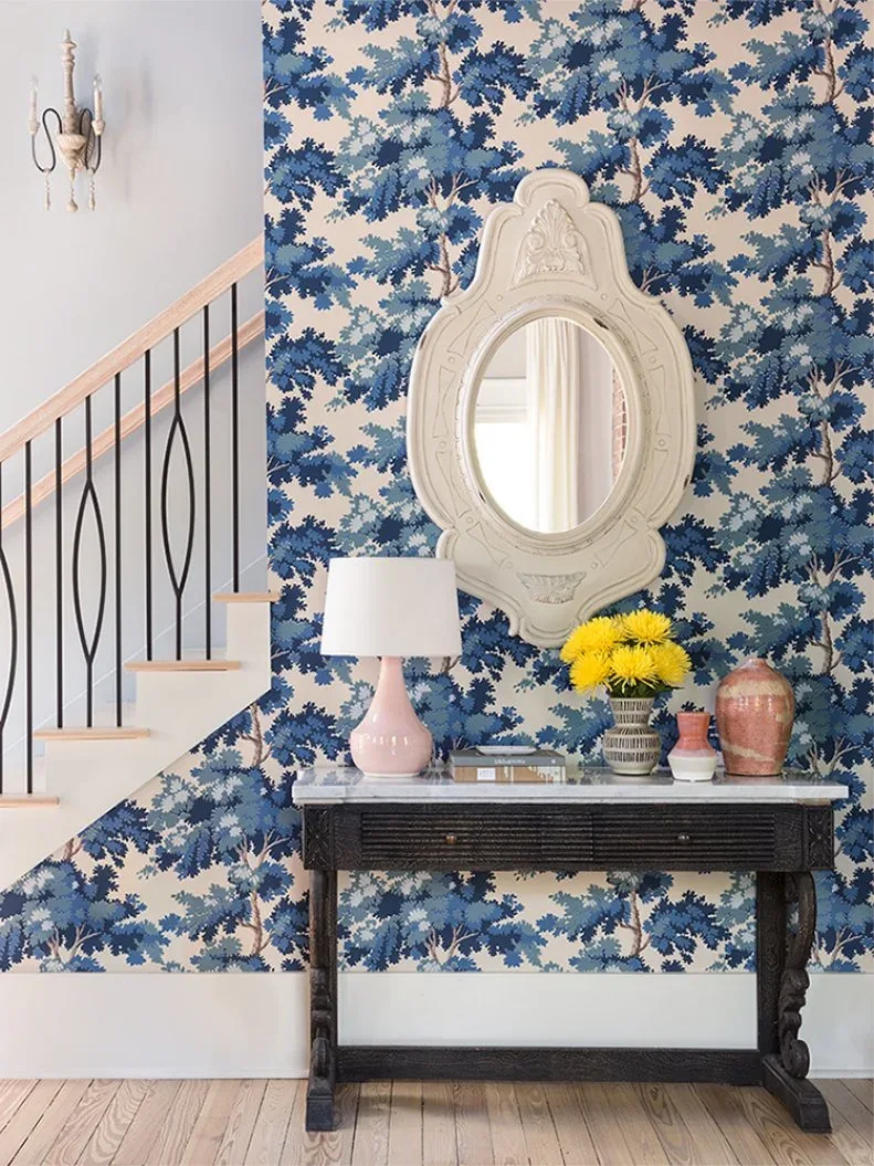 Black console beneath ornate pale oval mirror on blue leaf-pattern wallpaper beside stairs
