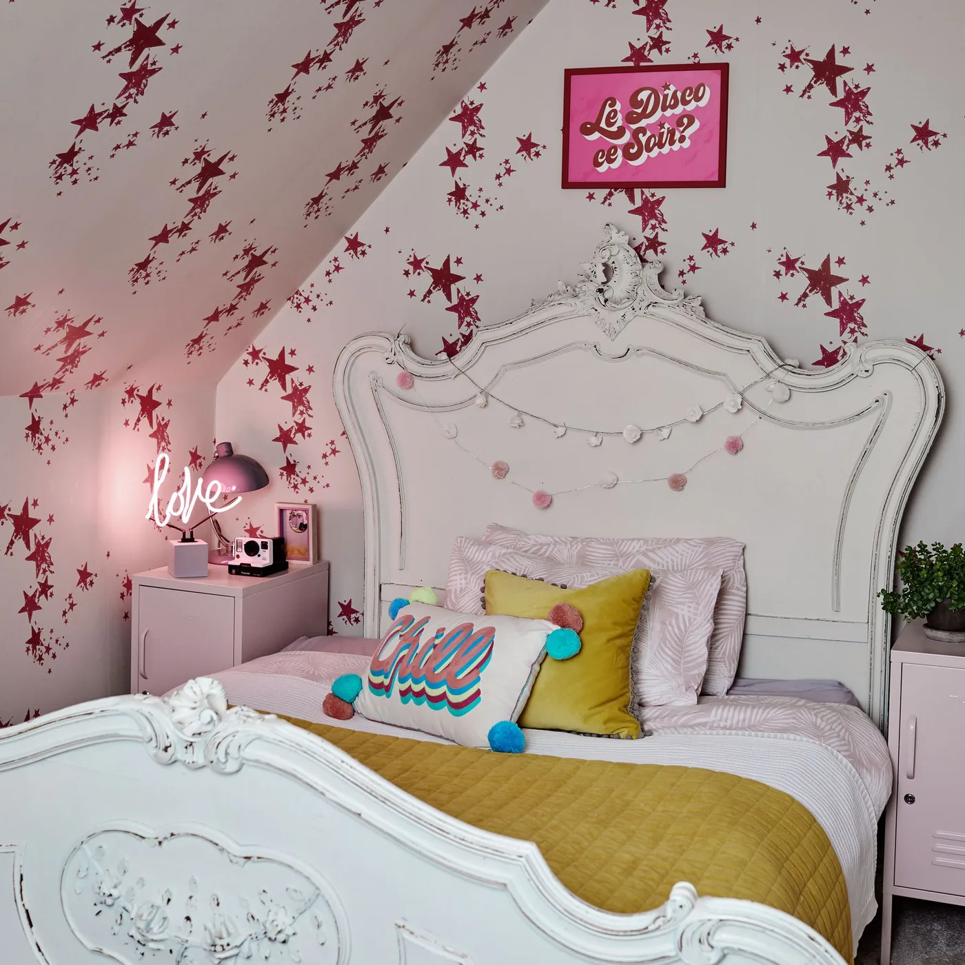 White ornate bed beneath pink star wallpaper extending across a sloped wall, with colorful cushions and pale bedside cabinets.