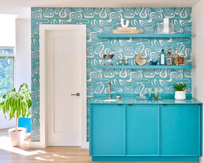 Turquoise floating shelves with glasses and bottles above matching cabinetry against blue patterned wallpaper