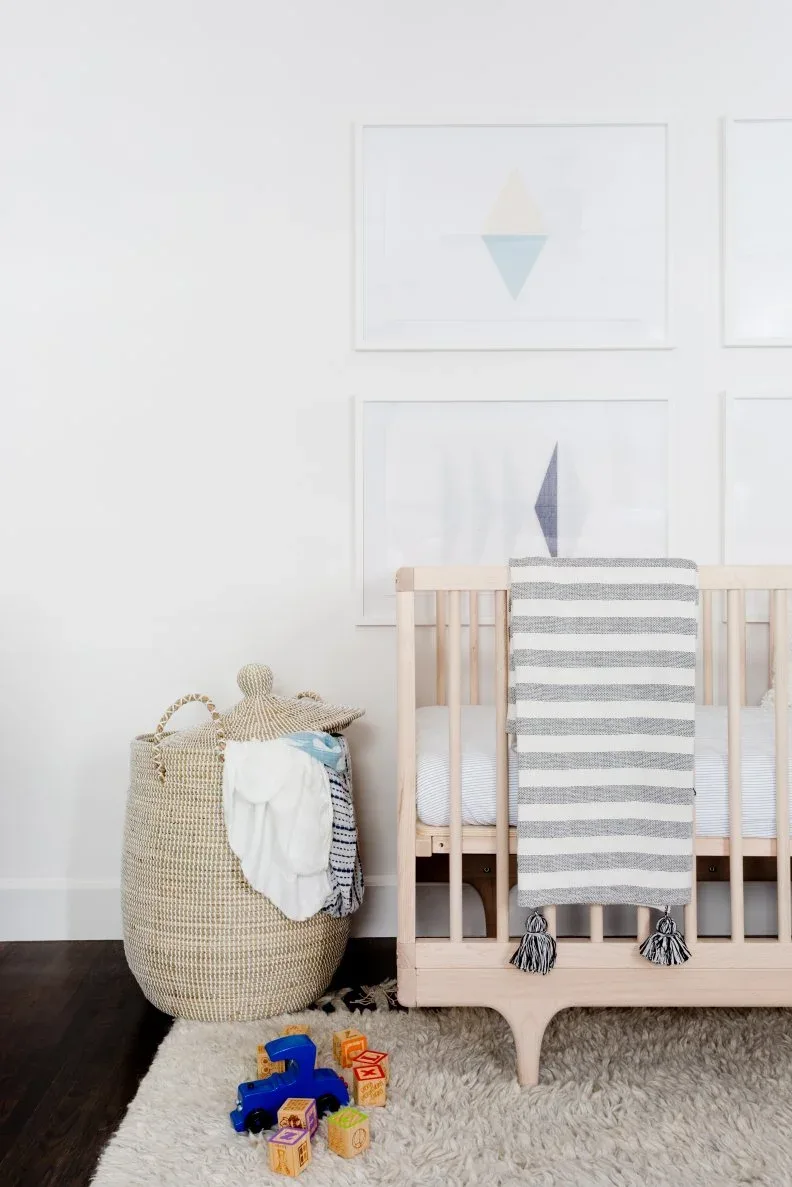 Pale wooden crib with draped gray-and-white striped throw beside woven basket beneath restrained framed artwork.