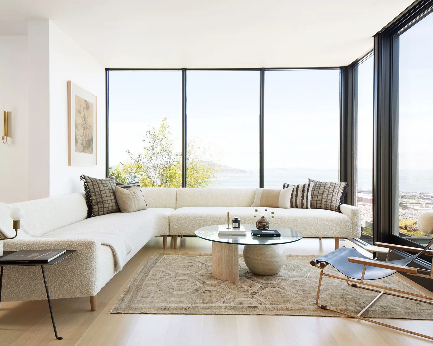 Pale curved sectional with wood-framed lounge chairs, round glass table and light wood floor beside broad windows