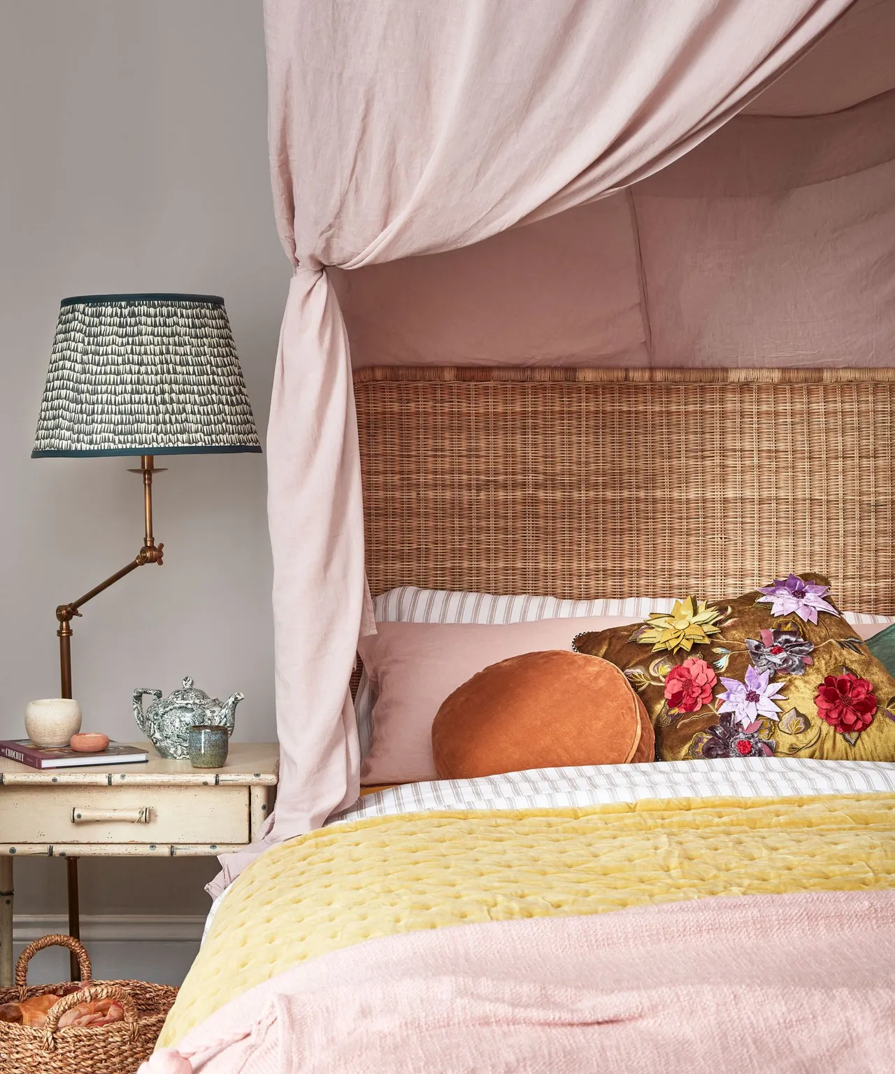 Bed with woven headboard, pink fabric canopy, pink and yellow bedding, embroidered cushions, small pale bedside drawer and green-shaded lamp.