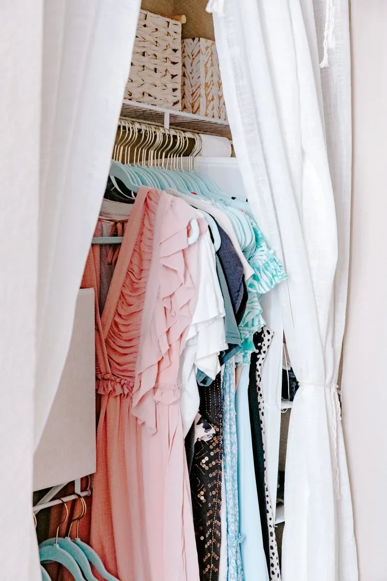 Student closet with pale curtain, coordinated hangers, pink and blue clothing and patterned storage boxes on an upper shelf.