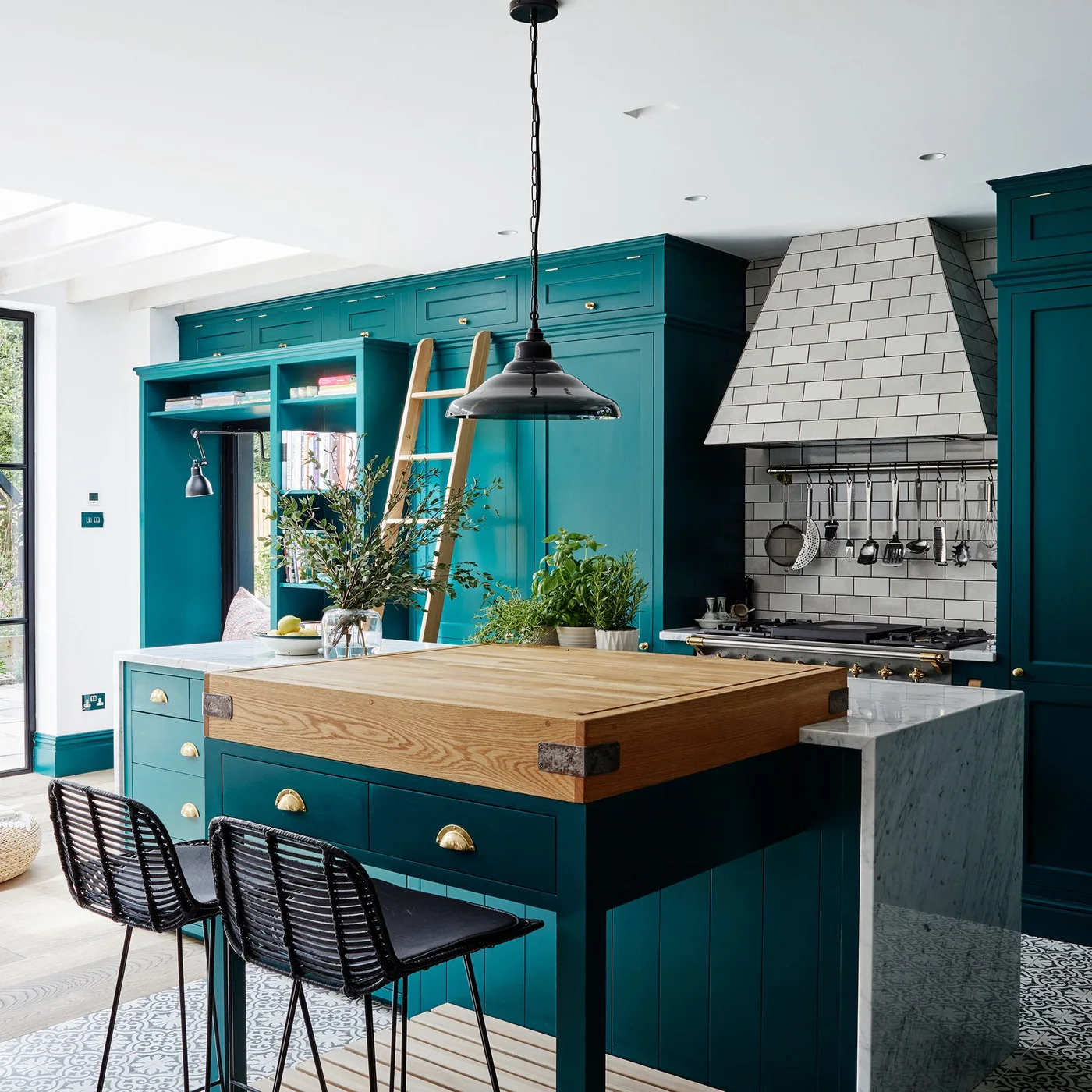 Deep teal kitchen cabinets surround a wooden-topped island beside dark stools, a pale tiled hood, open storage and a cabinet ladder