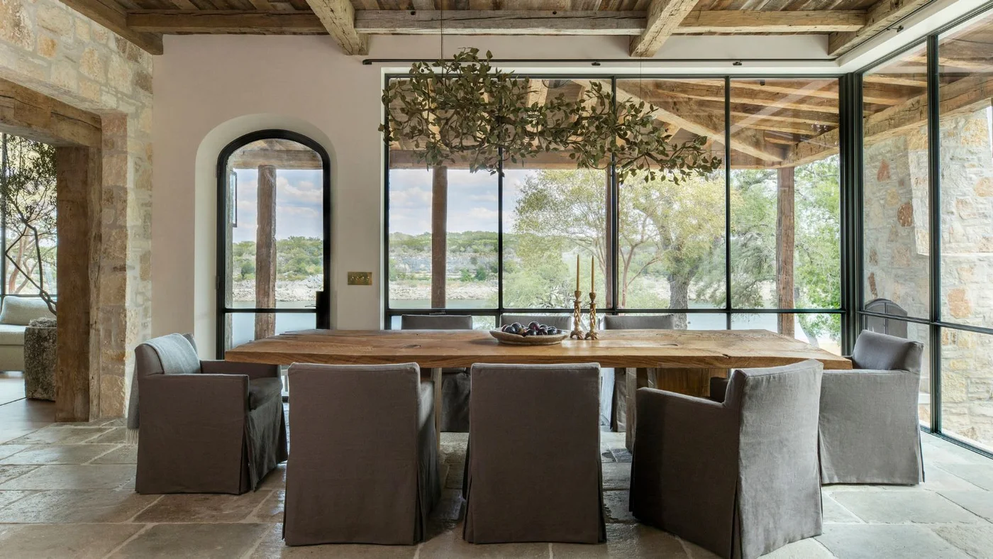A plain wood dining table and gray skirted chairs stand beside pale stone-textured walls, wood ceiling beams, broad black-framed glazing and a leafy overhead light