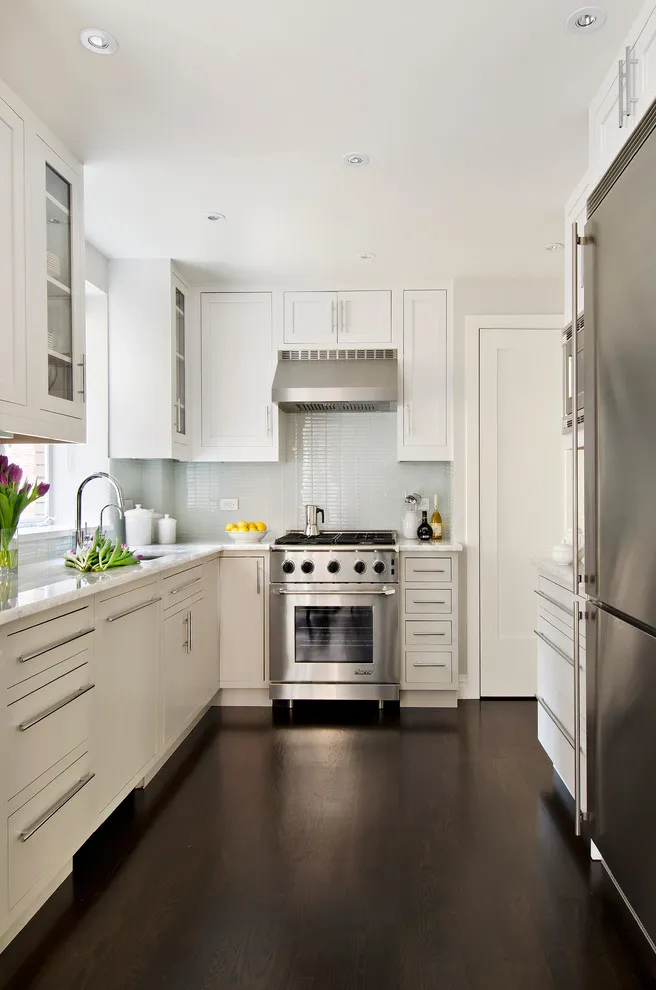 White cabinets line a narrow kitchen with a dark floor and bright sink window