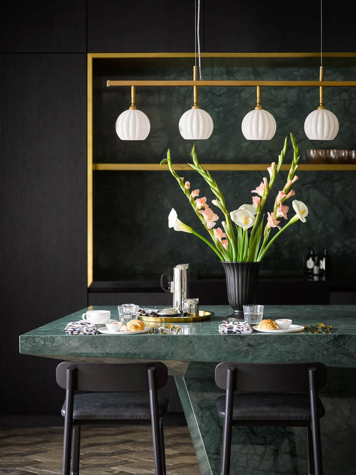 Four pale fluted pendant shades hang above a green veined dining table with dark chairs against a dark wall and warm-framed shelving