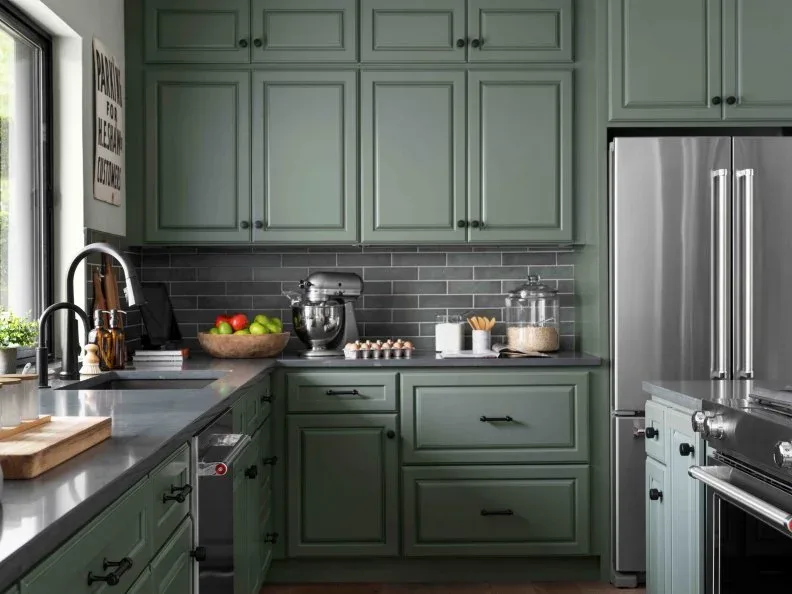 Muted green framed kitchen cabinets with charcoal rectangular backsplash tiles, light grout, gray countertop and a dark bridge faucet.