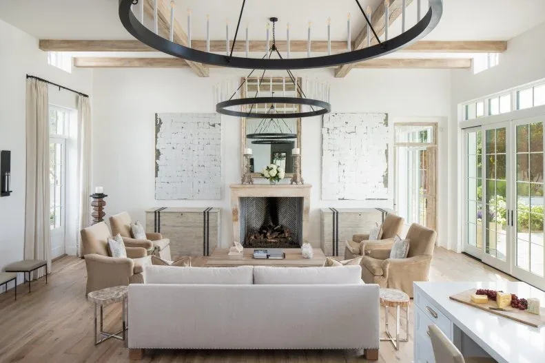 Pale fireplace beneath tall mirror and exposed beams in a light room with black ring chandeliers and grouped neutral seating
