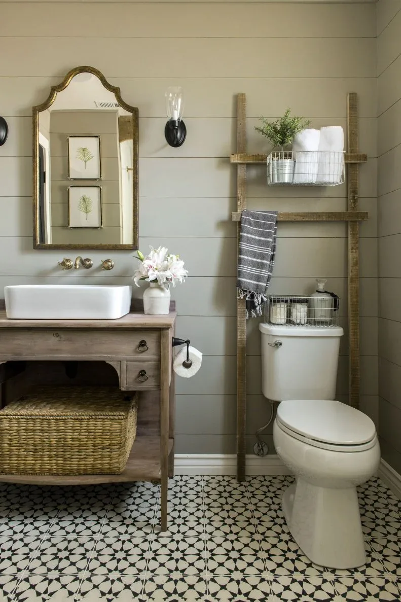 Powder room with pale horizontal wall panels, wooden vanity and woven basket, rectangular white basin, shaped golden mirror, ladder-style towel storage and white toilet.