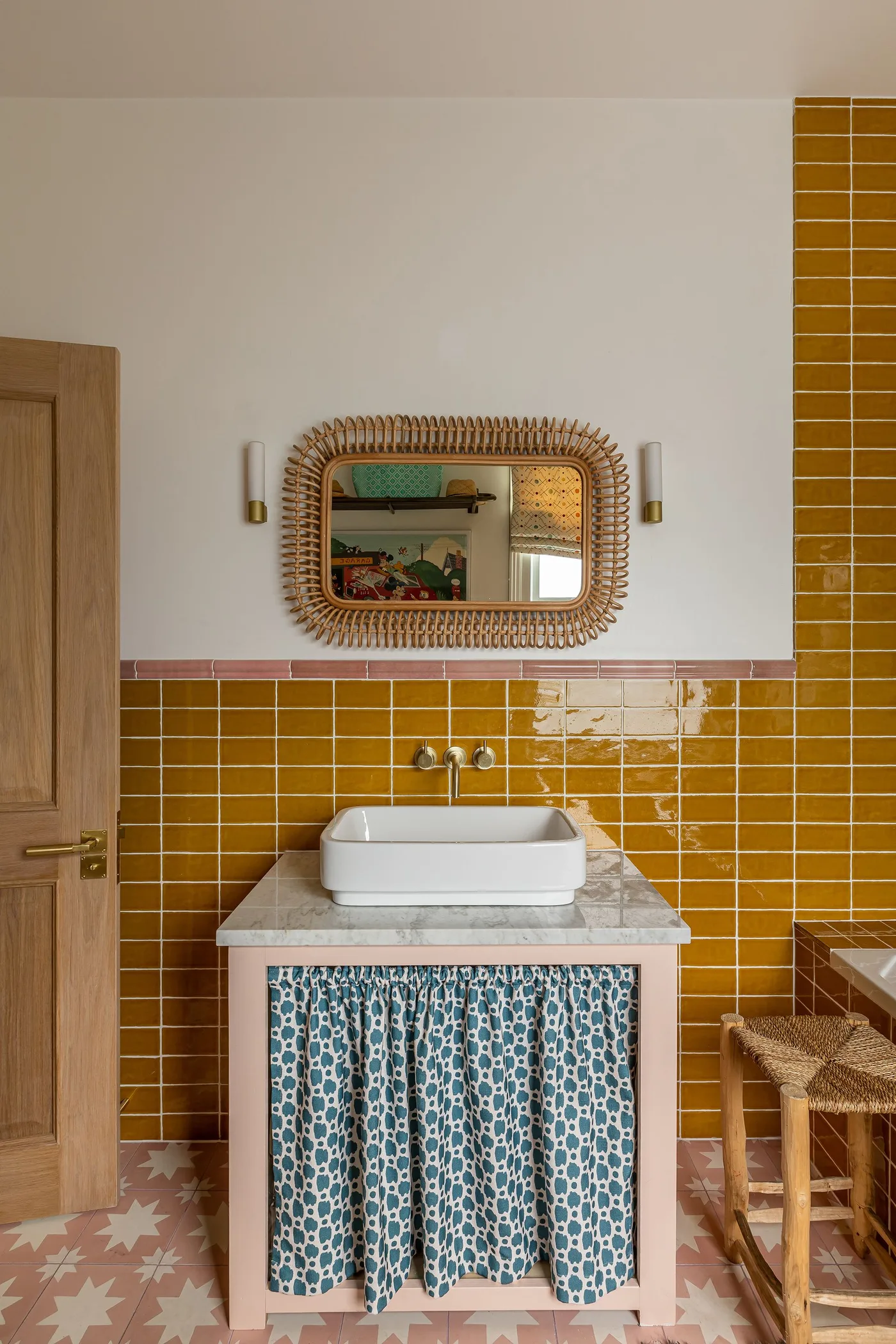 Country bathroom with mustard rectangular wall tiles, a white vessel basin, blue patterned sink skirt and woven rectangular mirror.