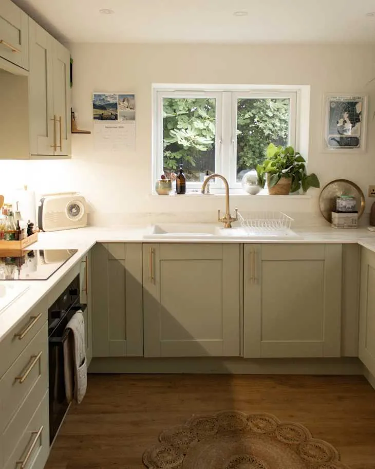 Sage-green kitchen cabinets with brass pulls, a white worktop, and a window over the sink