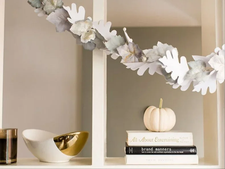 Pale and silver-toned leaf shapes form a light garland in front of open shelves holding a small pumpkin, books and a bowl