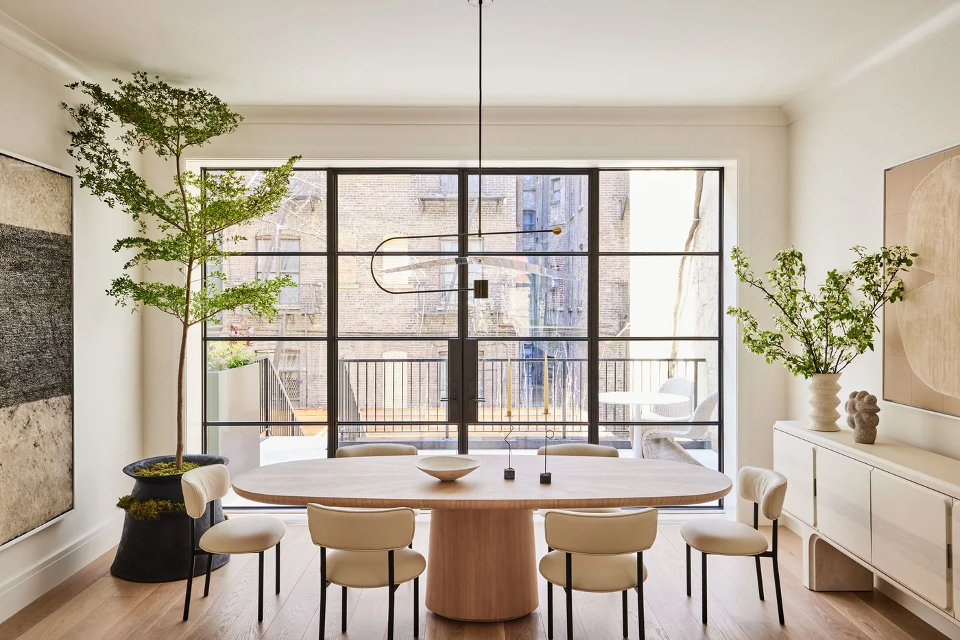 A pale oval wood dining table with a broad pedestal stands among cream chairs beside black-framed glazed doors, a pale sideboard and leafy plants