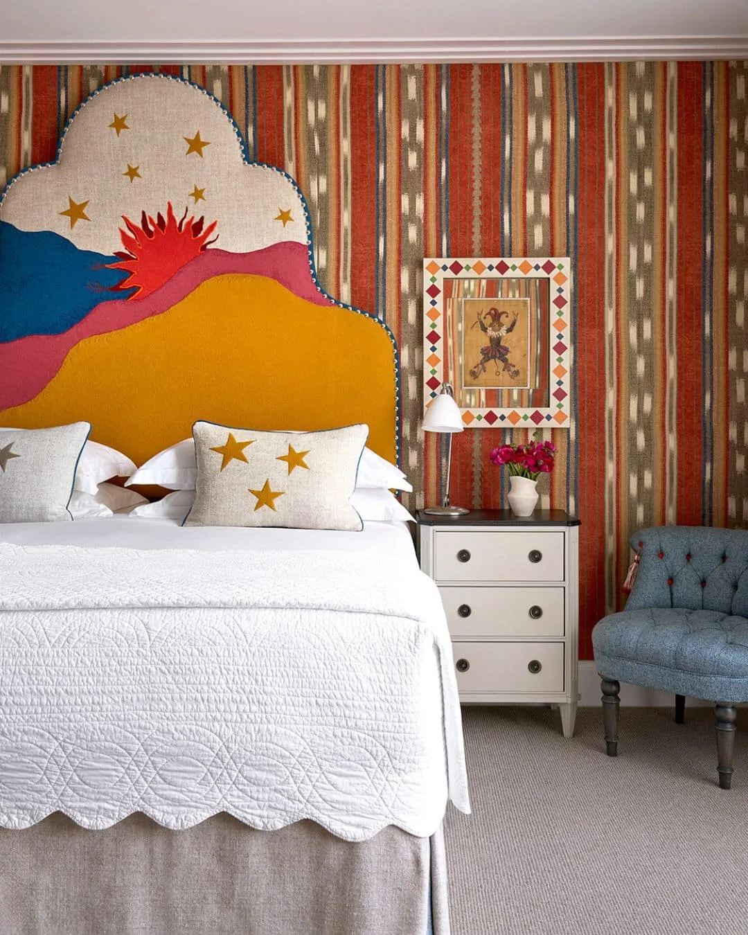 Tall pale headboard with a red sun and blue, pink and yellow hills against striped walls, with a star cushion.