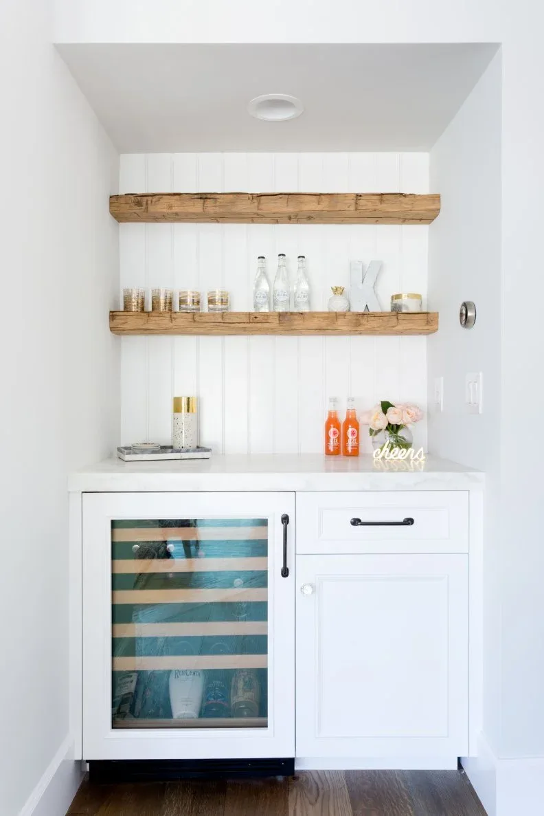 Two thick wood-toned floating shelves with bottles and glasses above a white cabinet and drinks fridge