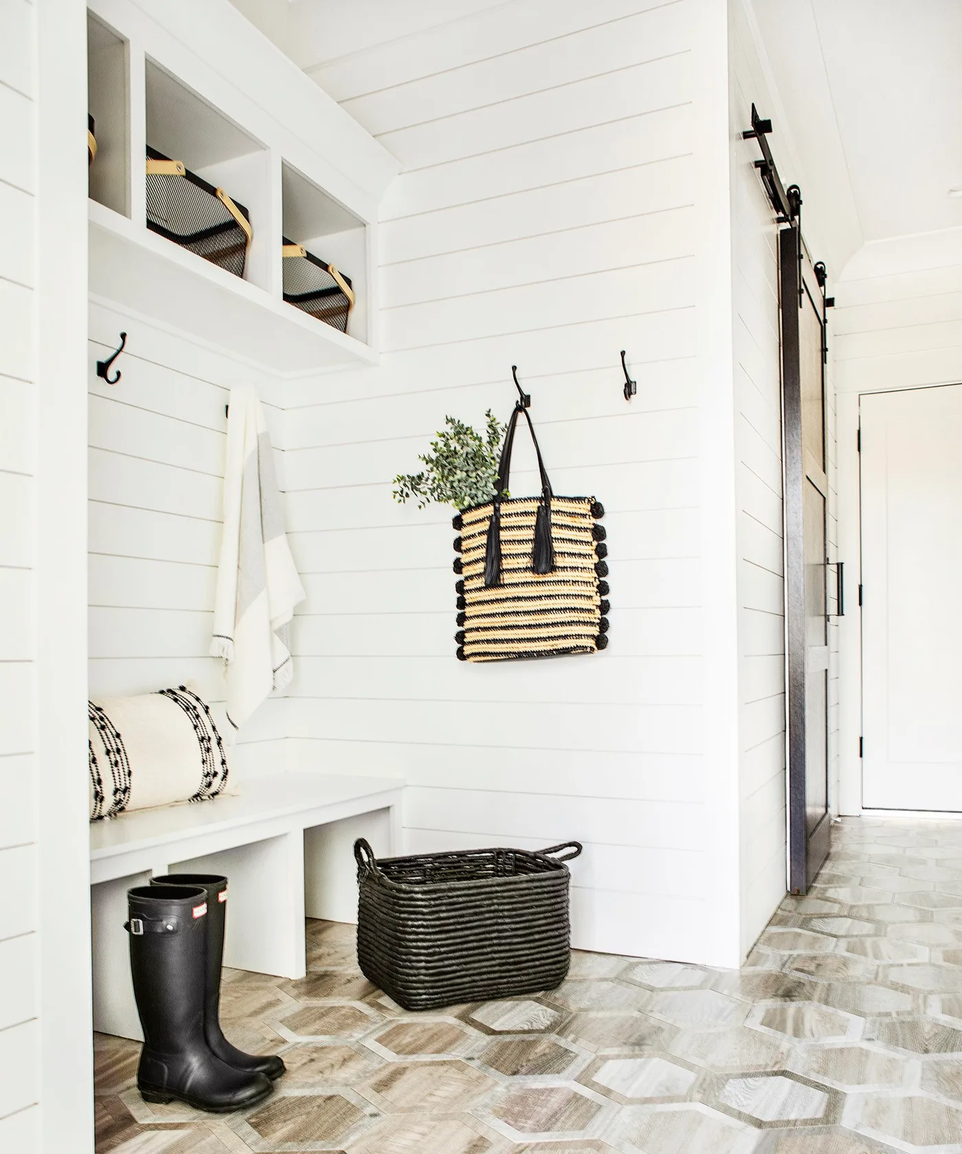 White paneled mudroom with a bench, upper cubbies, wall hooks, a striped bag, dark basket and tall black boots