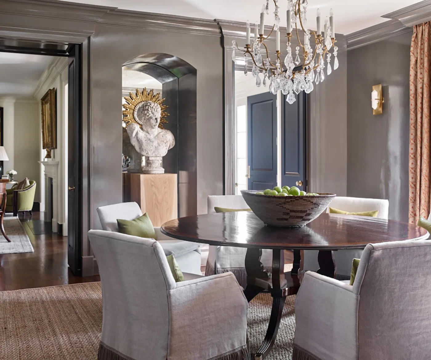 Round polished dark dining table with pale gray armchairs, glossy gray walls and a clear crystal chandelier.
