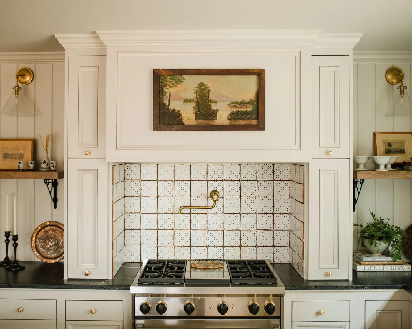 Cream kitchen cabinets and a decorative hood framing pale square backsplash tiles with small dark motifs behind the cooker.