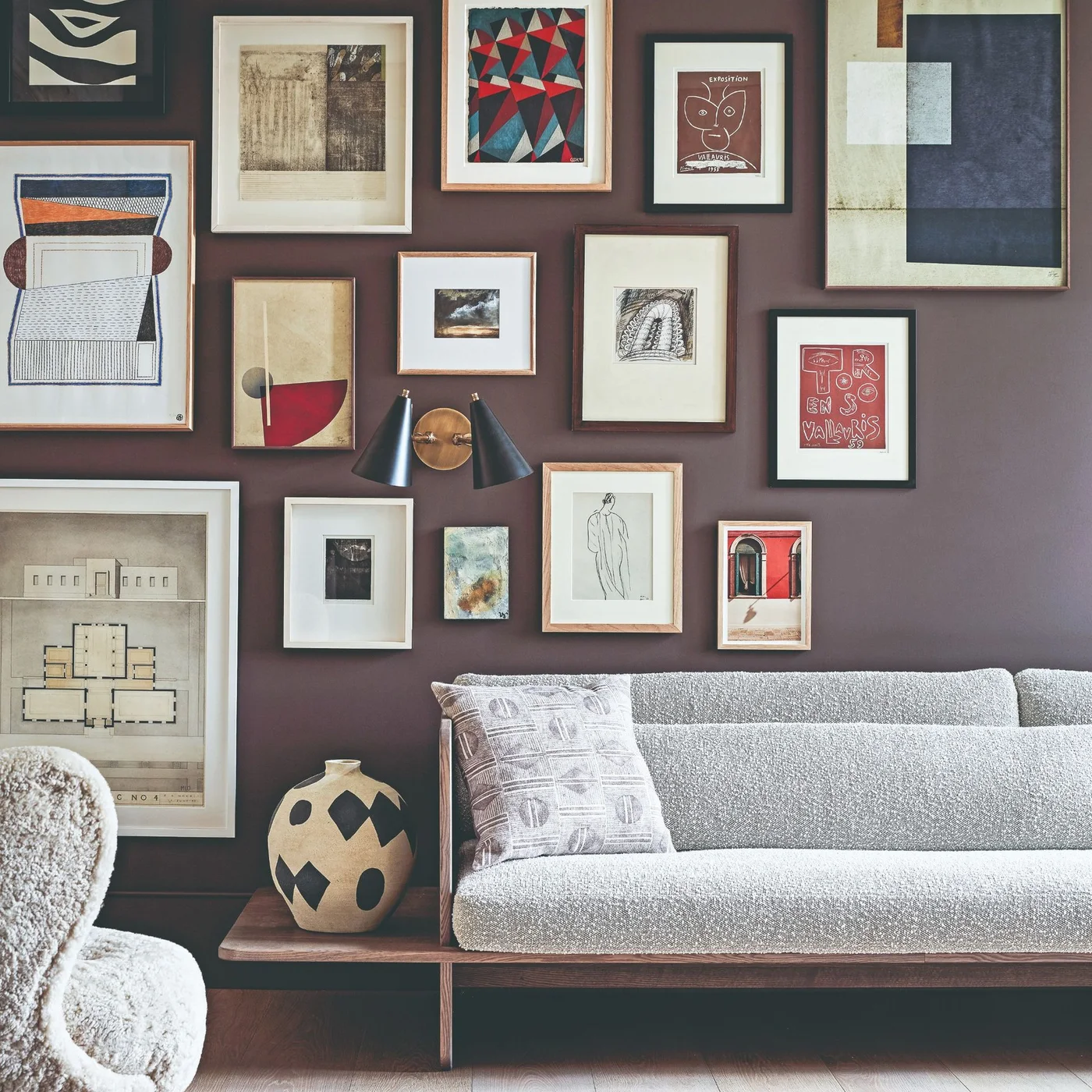 A low light gray sofa with wooden framing sits beneath a dense arrangement of mixed framed artwork on a muted plum wall