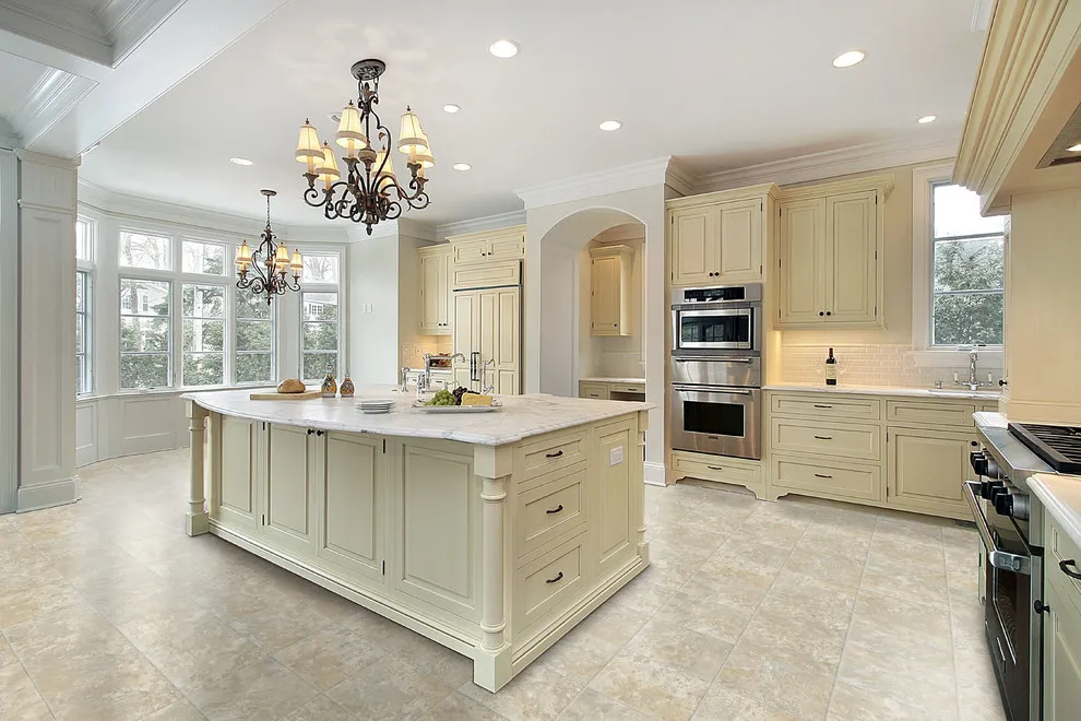 Cream cabinetry and a large pale-topped island sit beneath two decorative chandeliers in a bright kitchen