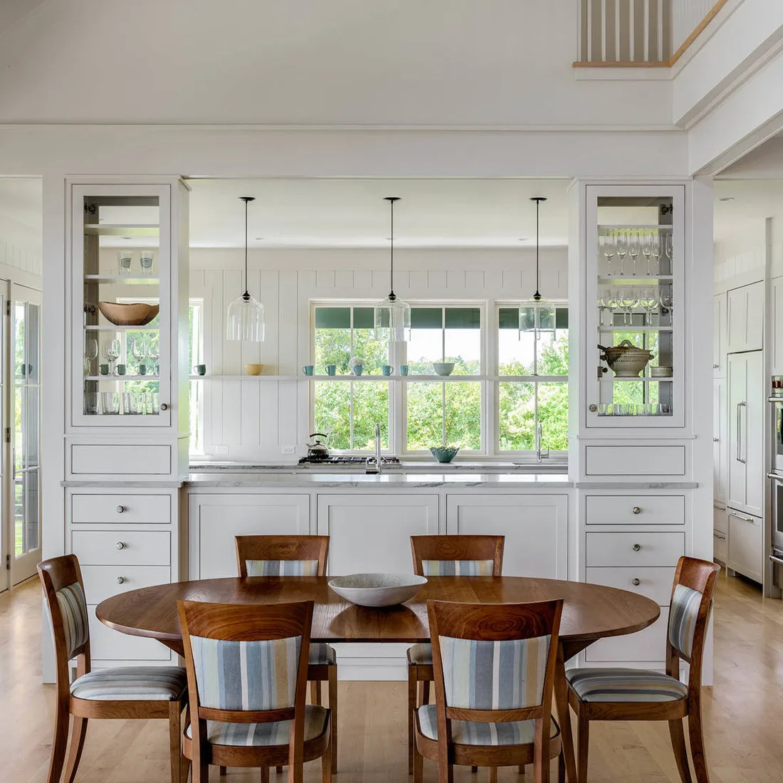 Wood oval dining table with striped-cushion chairs in front of white cabinets and a central window