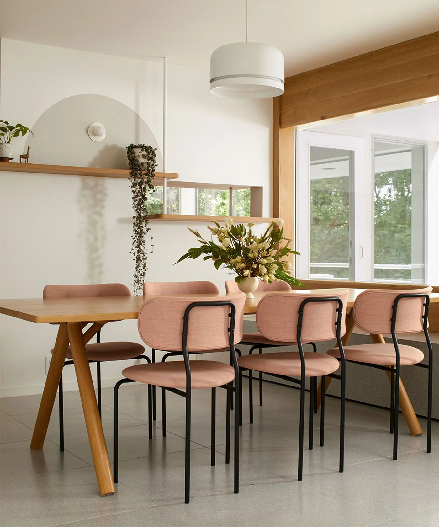 Light dining room with a long wood table, dusty pink chairs on black frames, white drum pendant and horizontal wall mirror
