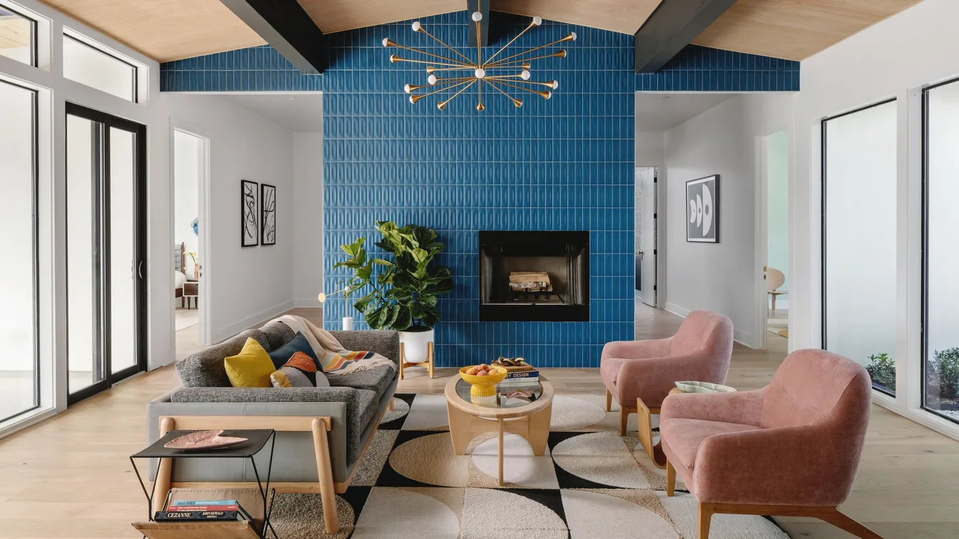 A blue tiled fireplace wall follows a pitched ceiling behind gray seating and pink chairs beneath black beams