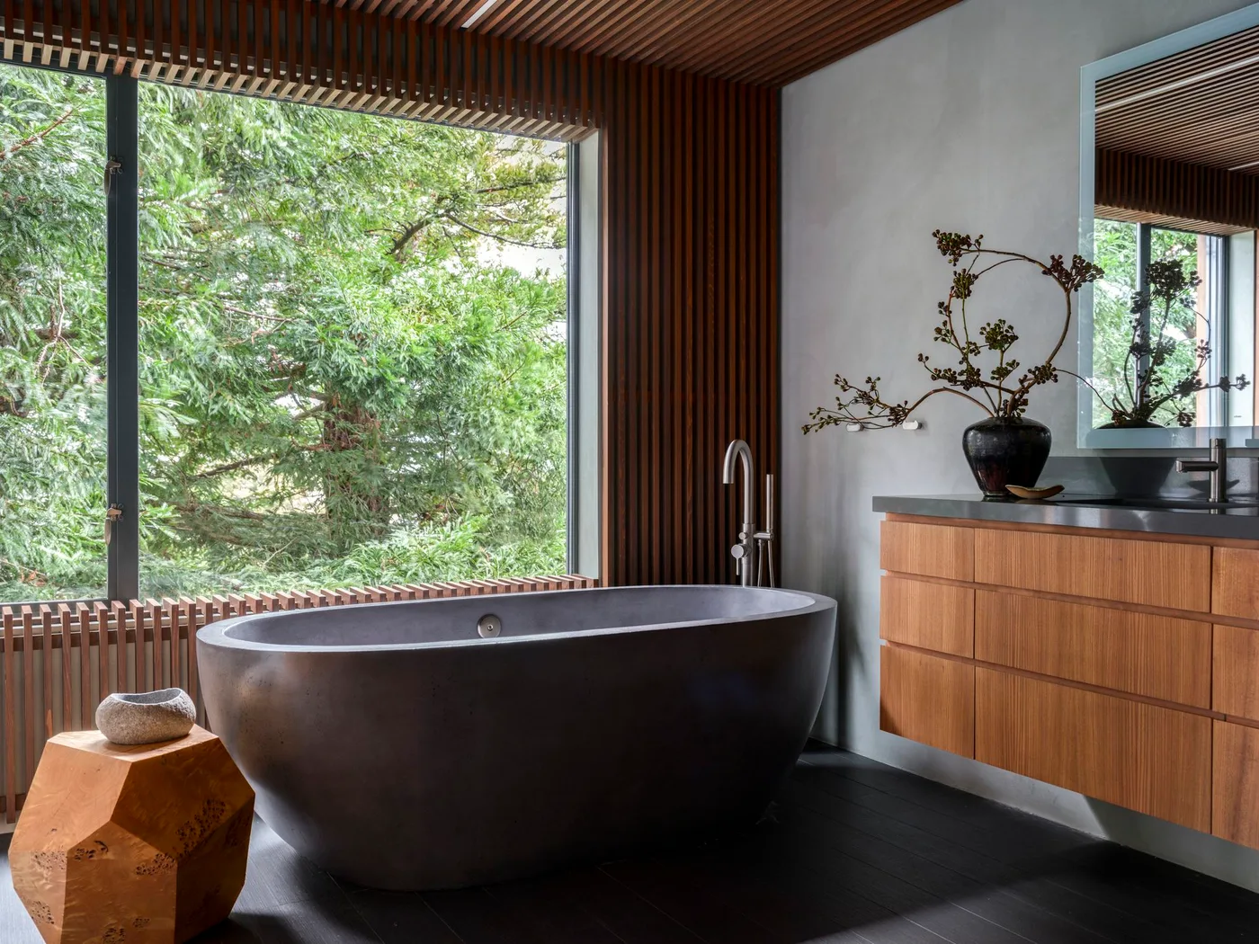 Dark oval bath beside warm wood slats, a broad garden window and a floating wood vanity