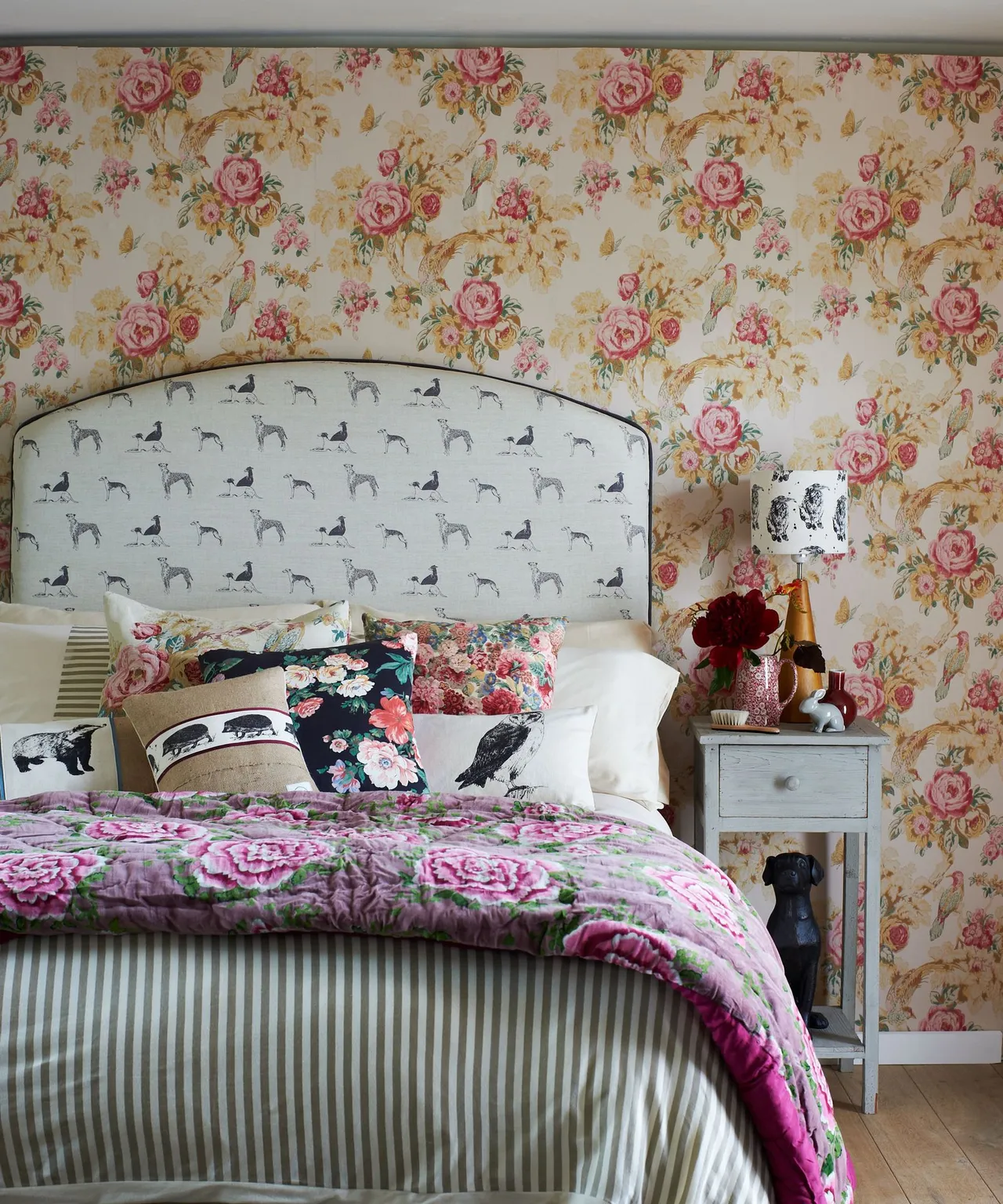 Bedroom with rose-patterned wallpaper, curved animal-print headboard, striped bed skirt, floral bedcover and several patterned cushions.