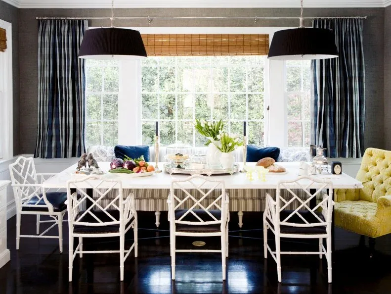 Warm textured shade above a broad dining window framed by dark curtains behind white chairs