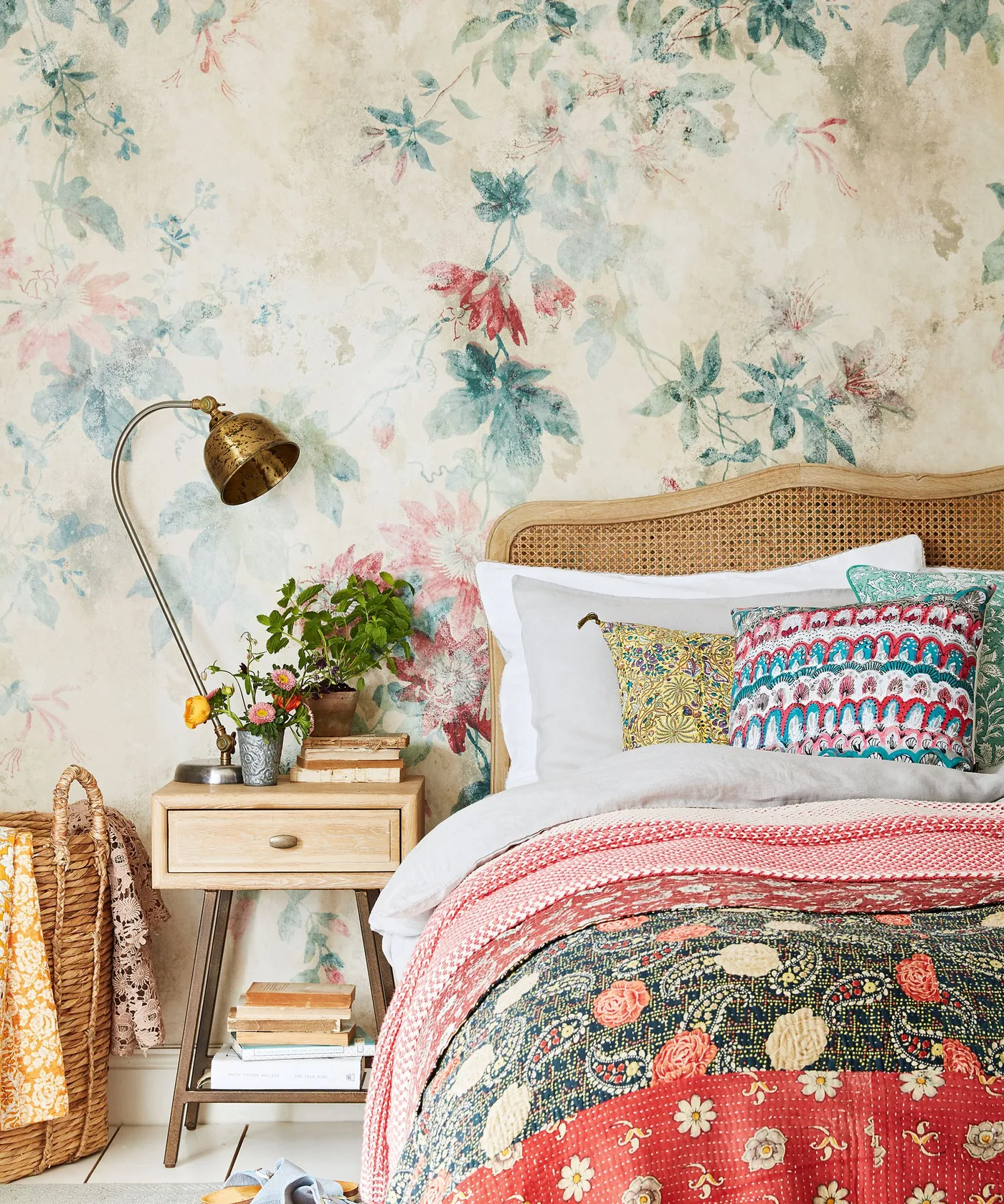 Bedroom detail with faded-looking floral wallpaper, curved woven headboard, mixed patterned pillows, floral bedcover, pale wooden nightstand and curved reading lamp.