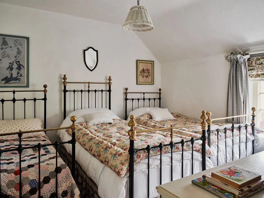 Shared cottage bedroom with three dark metal beds, gold-colored finials, pale walls, floral and patchwork quilts and a woven-looking pendant