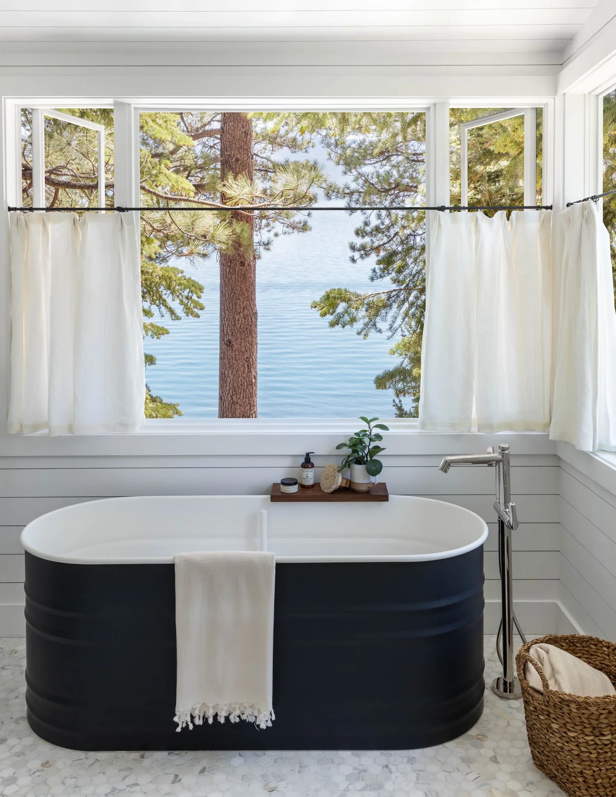 A dark freestanding tub with white interior stands beside white horizontal boards, broad windows with short curtains, a woven basket and a small wood bath tray