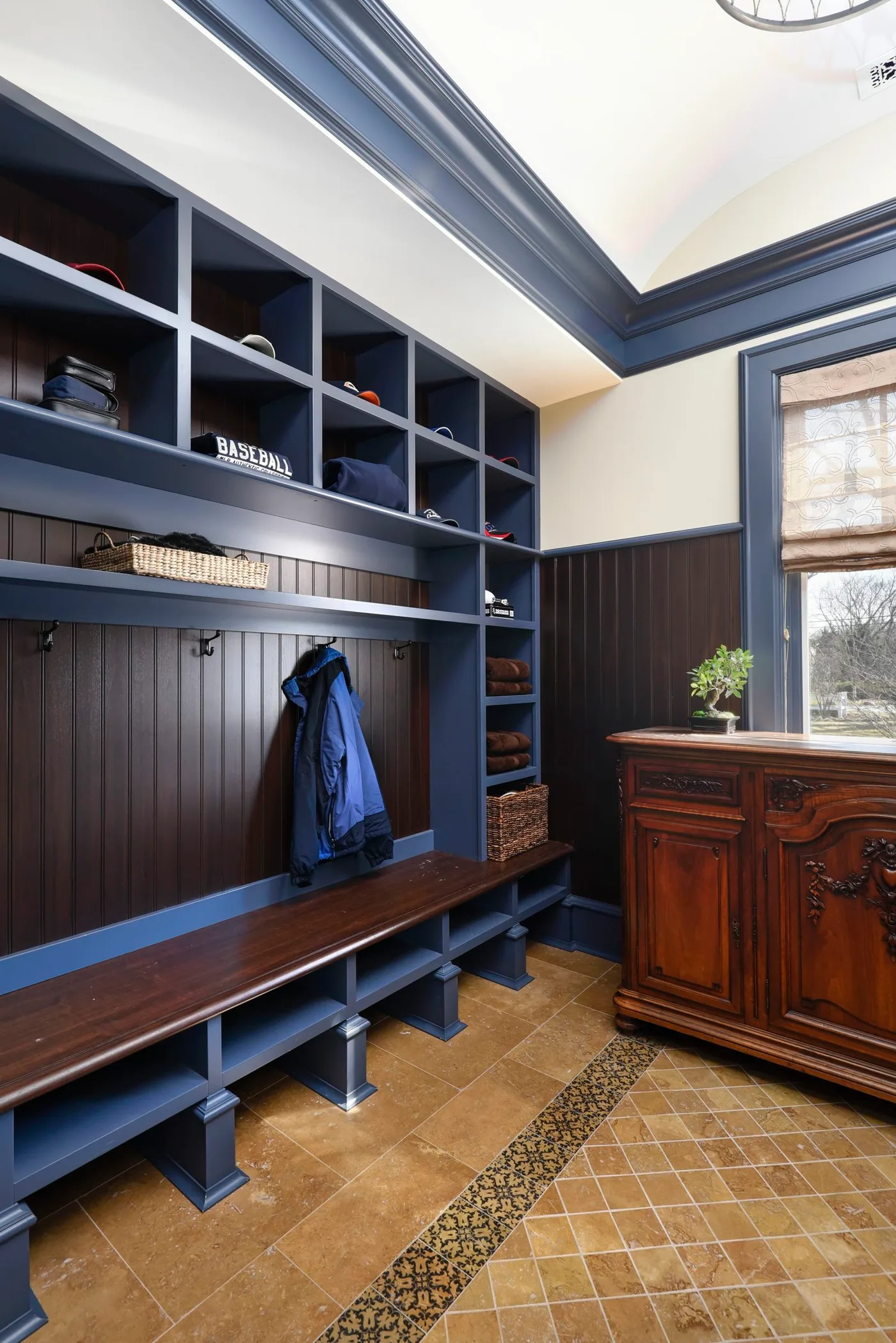 Deep blue mudroom lockers with dark brown paneled backing and bench, upper cubbies, shoe shelves, a wooden sideboard, window, and warm-toned floor.