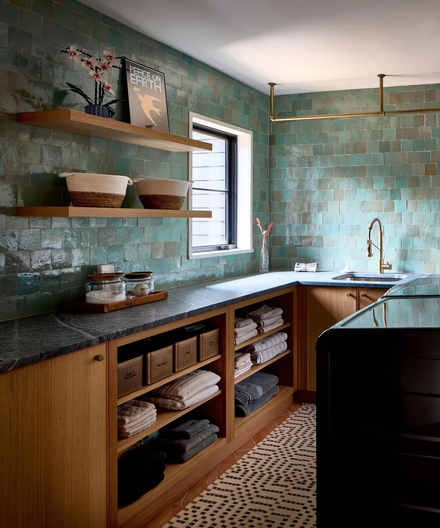 Green tiled utility walls with dark counter, wood cupboards and open towel shelves beneath a hanging rail