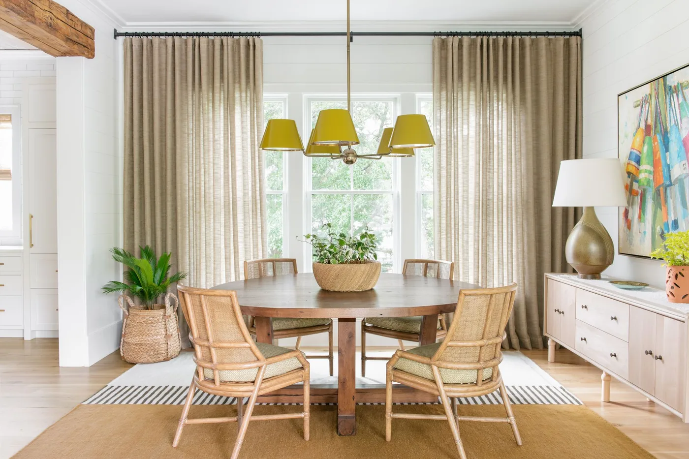 Pale dining cabinet with drawers beside a round wood table, woven chairs and yellow lamp shades