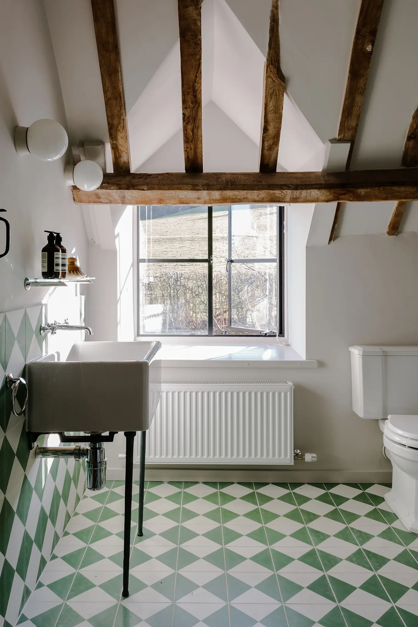 Bathroom with exposed ceiling beams, green and pale geometric floor tiles, a gray basin on dark legs and a white toilet.