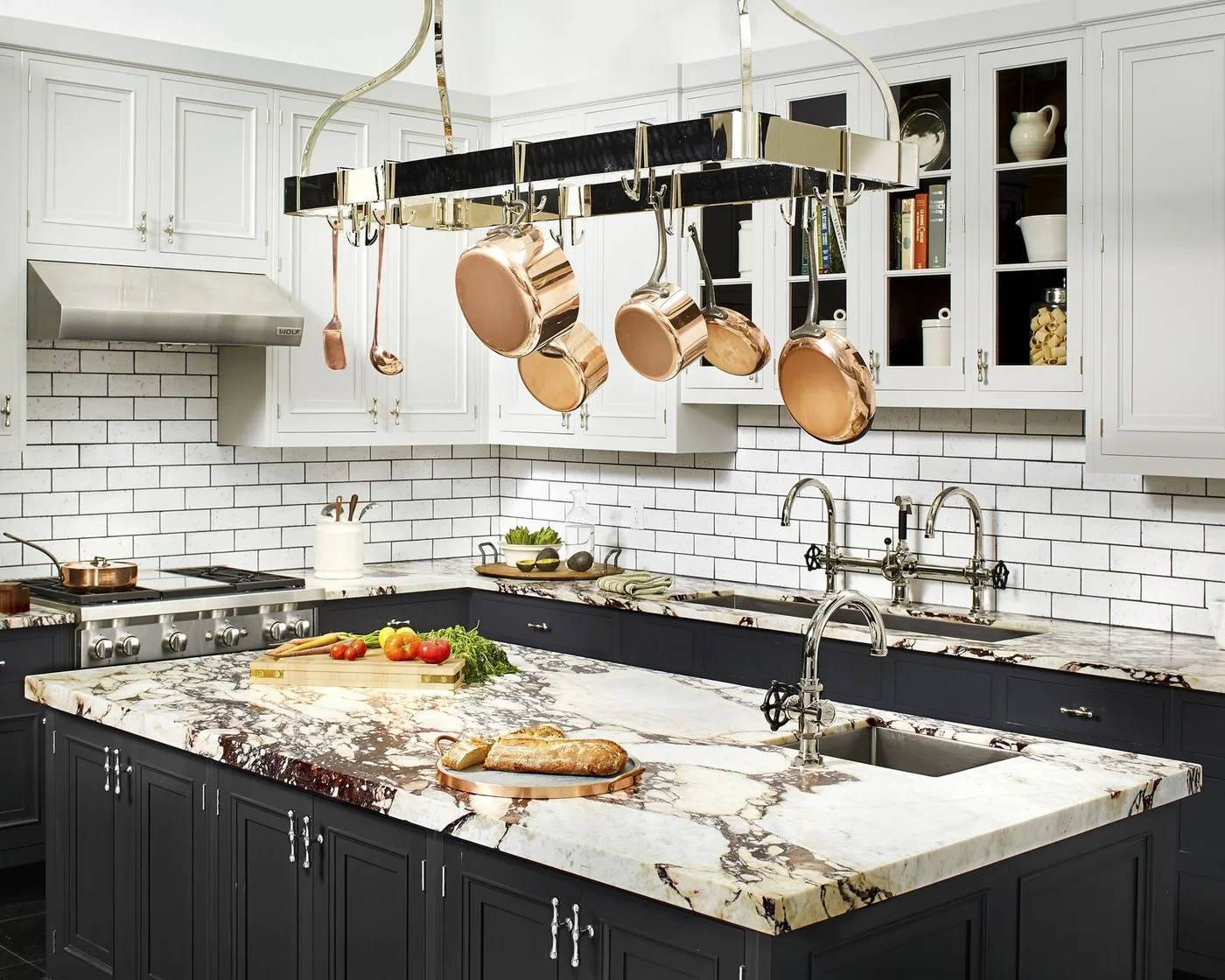 A kitchen has dark lower cabinets and an island below white upper cupboards, white subway tile, a hanging pan rack and strongly veined pale counters