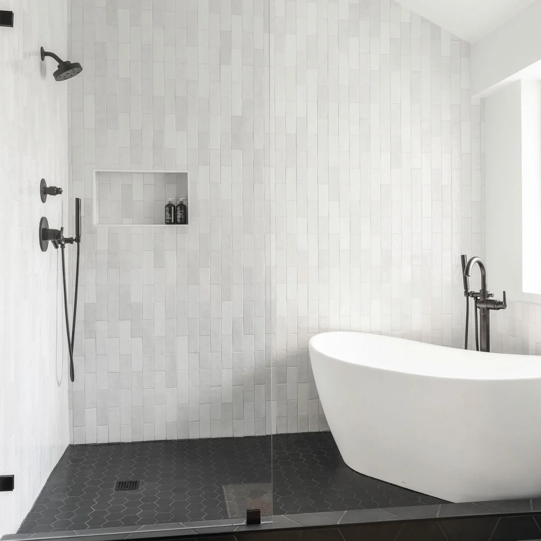 White vertical shower tiles and a long niche sit above a dark hexagonal floor beside a freestanding white tub