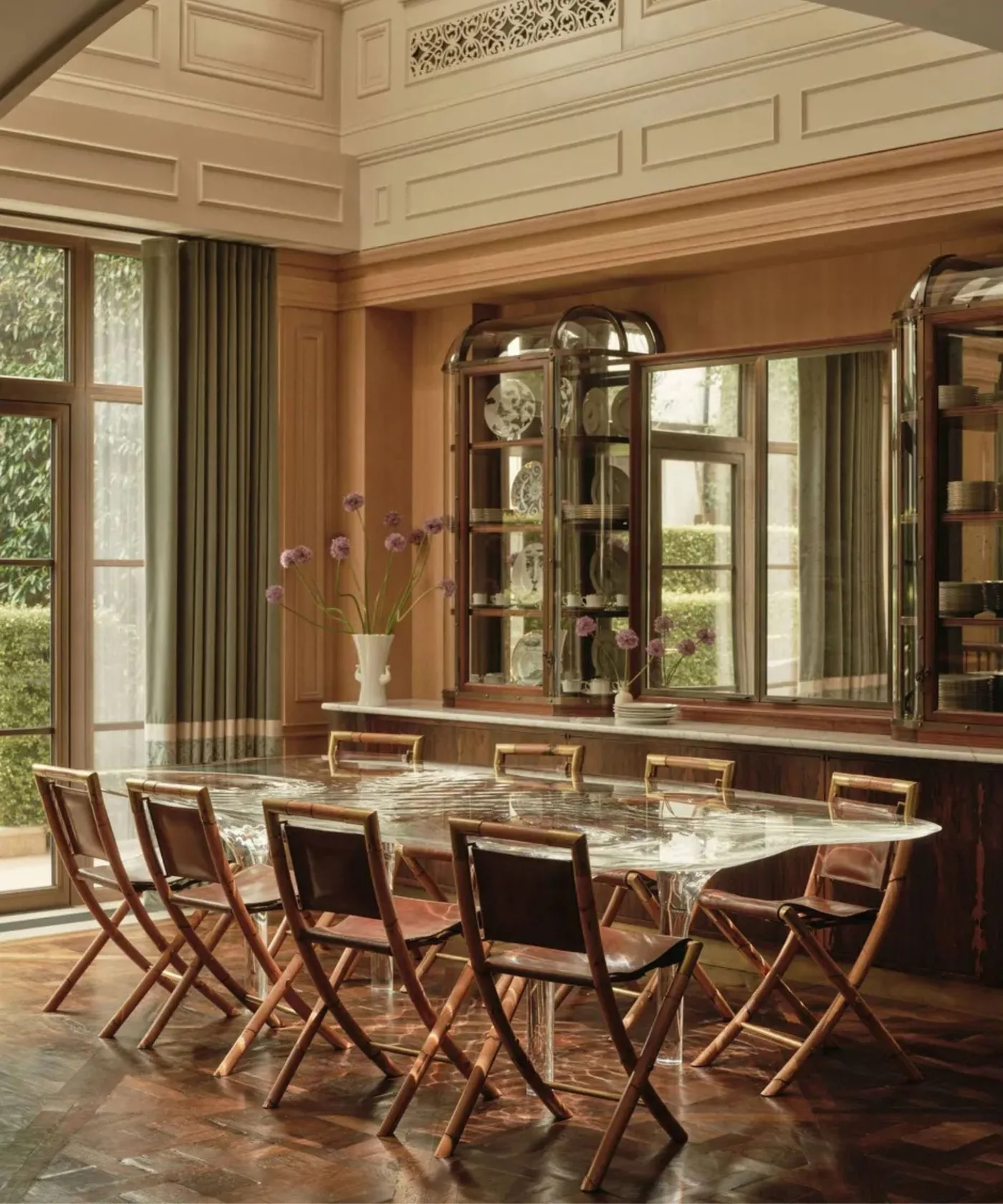 A transparent dining table and wooden chairs stand beside tall glazed cabinets beneath pale wall paneling and curtains